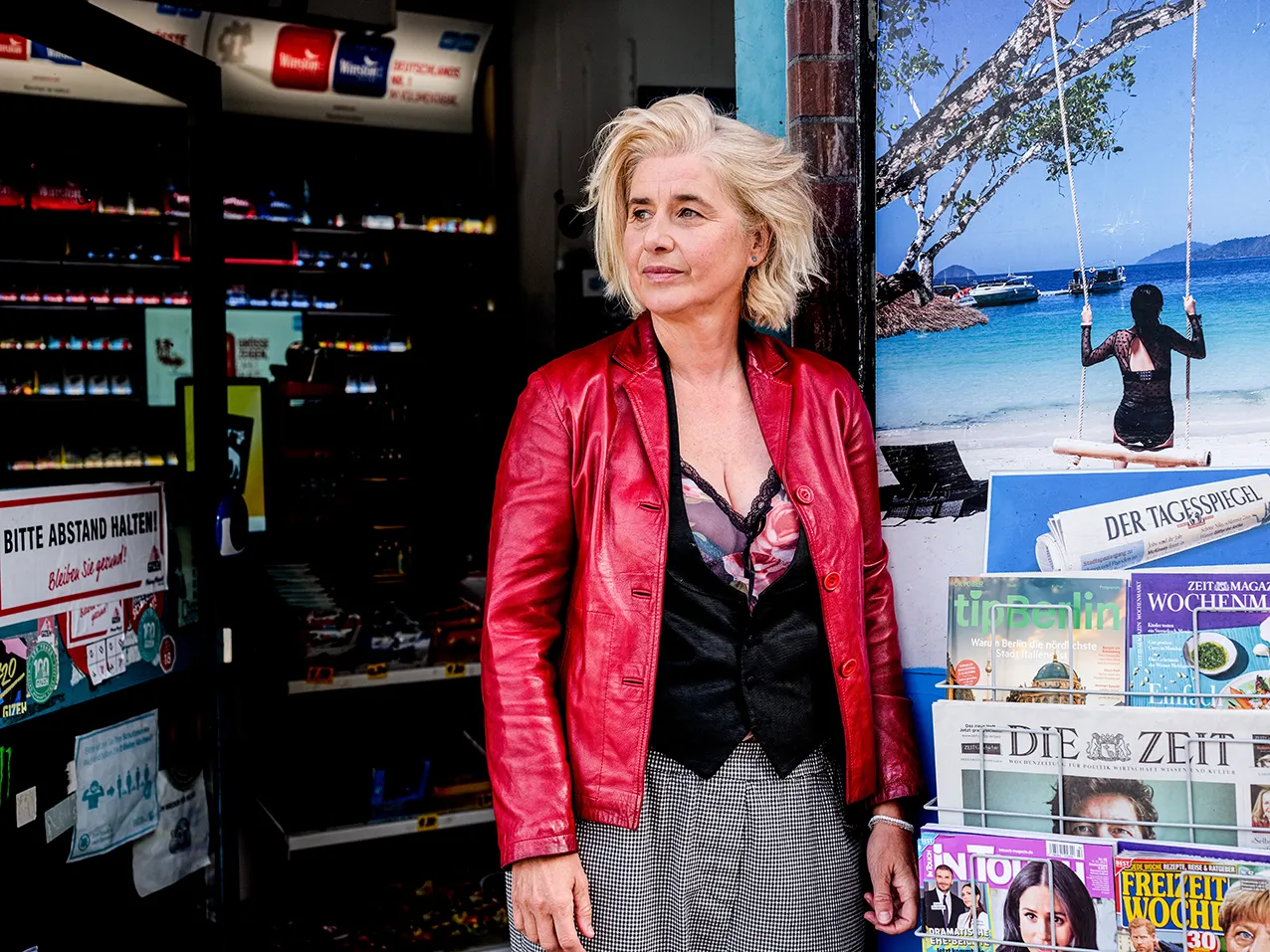 Angelika Fink in roter Lederjacke vor einer Trafik, Zeitschriften und Regale im Bild