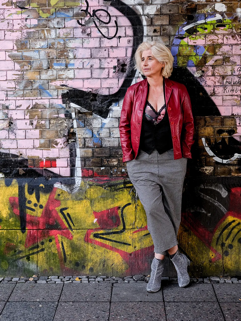 Angelika Fink in Ganzfigur, rote Lederjacke, vor einer bunt besprühten Ziegelwand