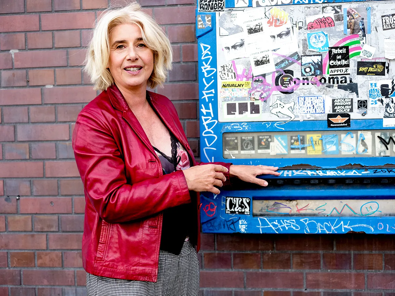 Angelika Fink in roter Lederjacke an einem mit Aufklebern beklebten Automaten